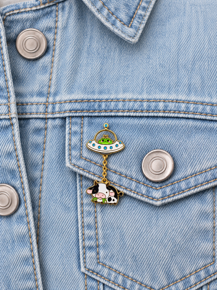 Alien Cow UFO Dangle Pin displayed on a light-wash denim jacket beside a metal button