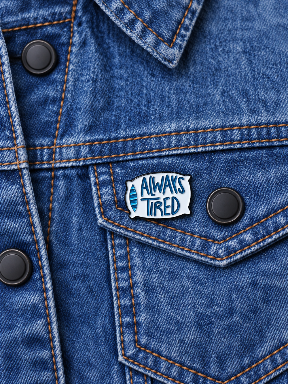 Always Tired Pillow Pin displayed on an acid-wash denim jacket beside a metal button