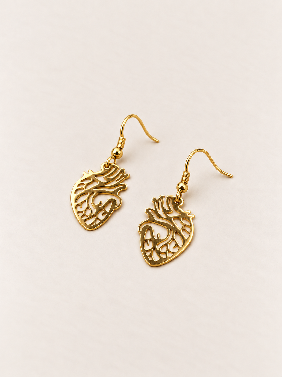 Anatomical Heart Drop Earrings in gold lying flat on a warm neutral surface