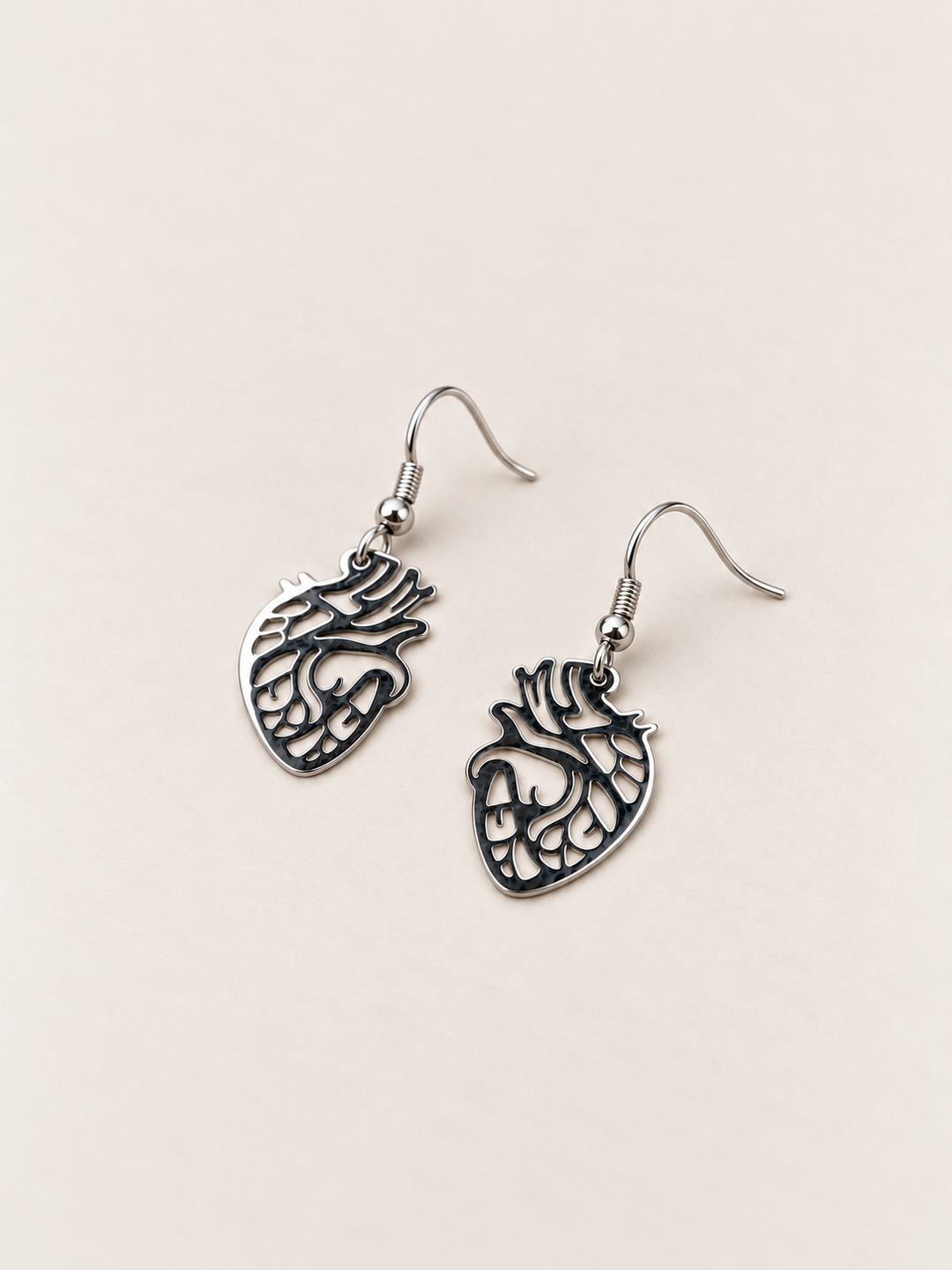 Anatomical Heart Drop Earrings in silver lying flat on a warm neutral surface