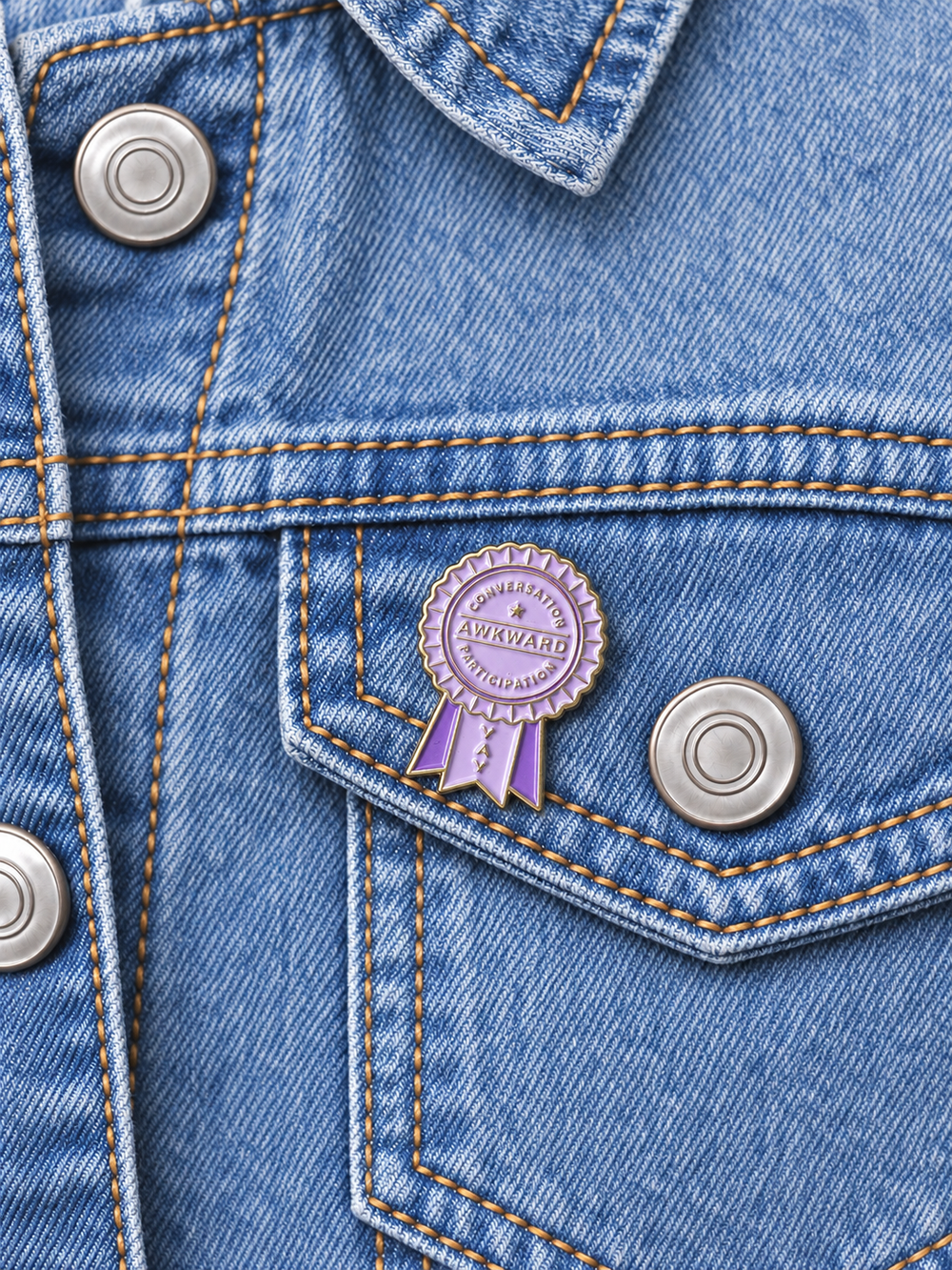 Awkward Conversation Participation Ribbon Pin displayed on a light-wash denim jacket beside a metal button