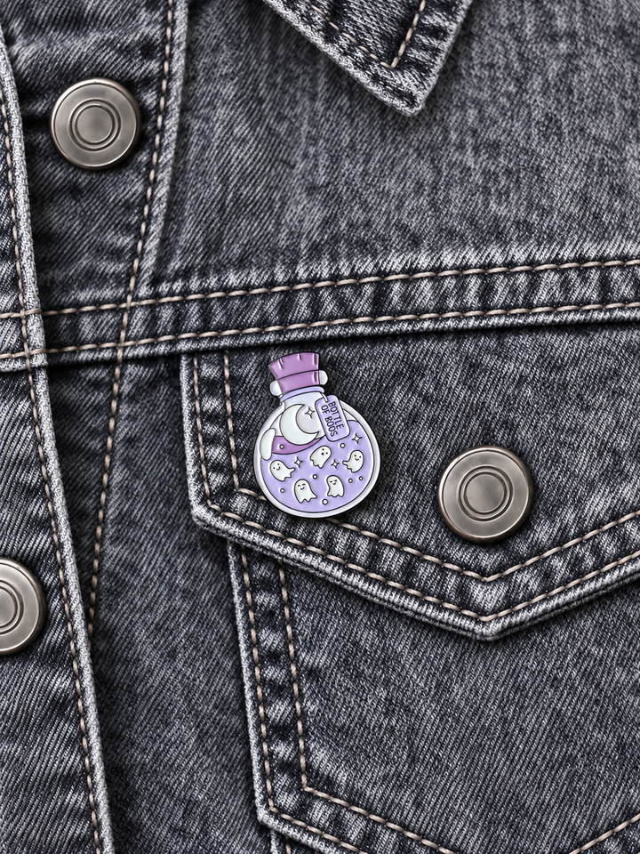 Bottle of Boos Ghost Potion Pin displayed on a gray acid-wash denim jacket beside a metal button