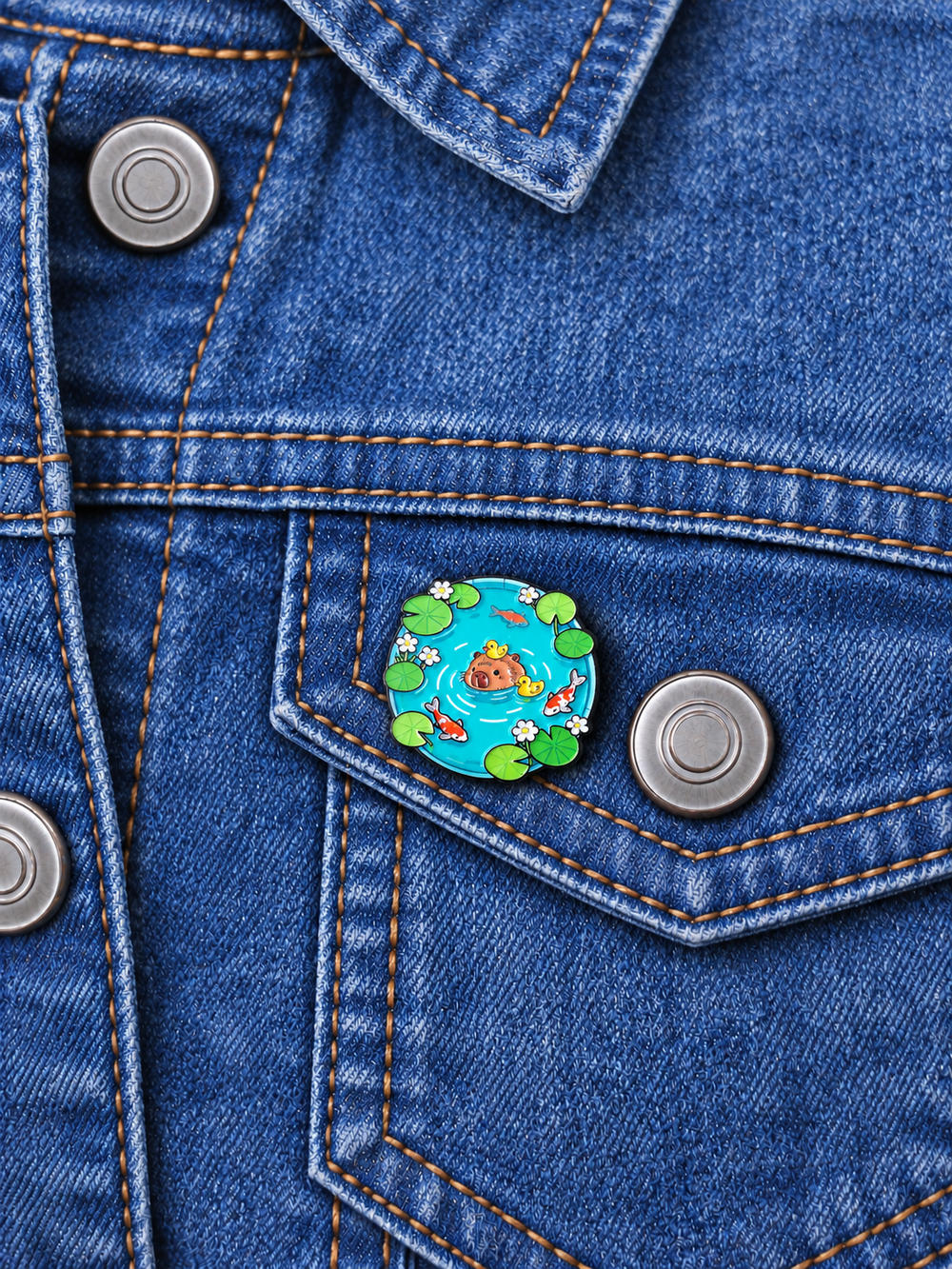 Capybara Koi Pond Pin displayed on a medium-blue denim jacket beside a metal button