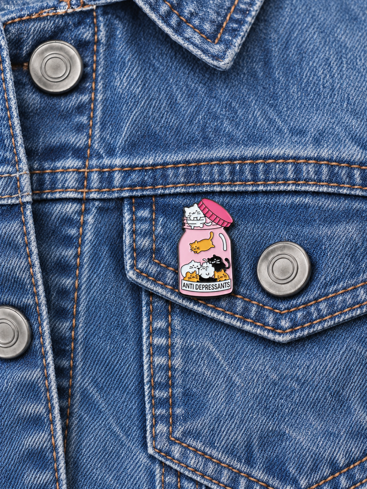 Cats Antidepressants Bottle Pin displayed on a blue denim jacket beside a metal button