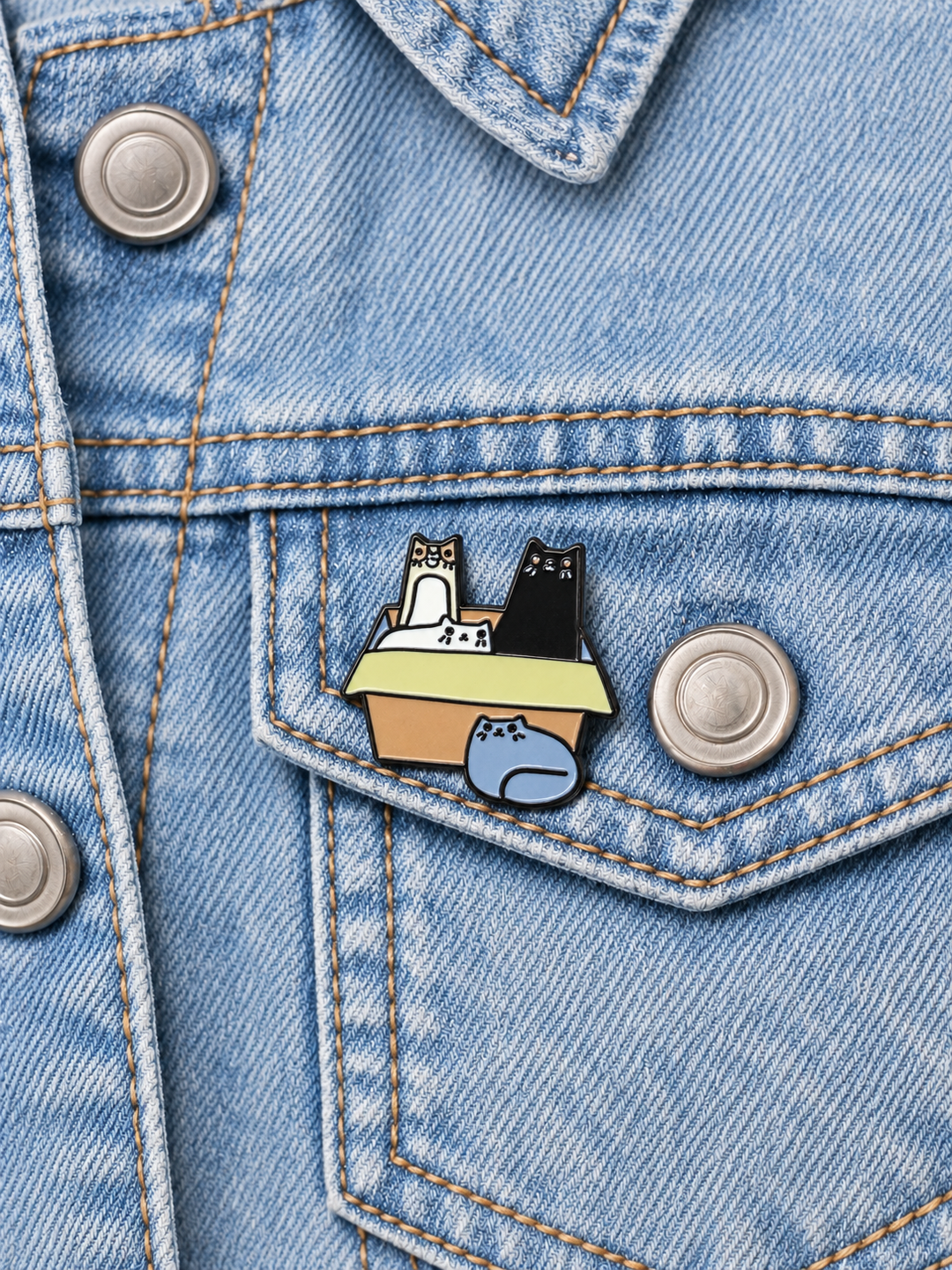 Cats in Cardboard Box Pin displayed on a blue denim jacket beside a metal button