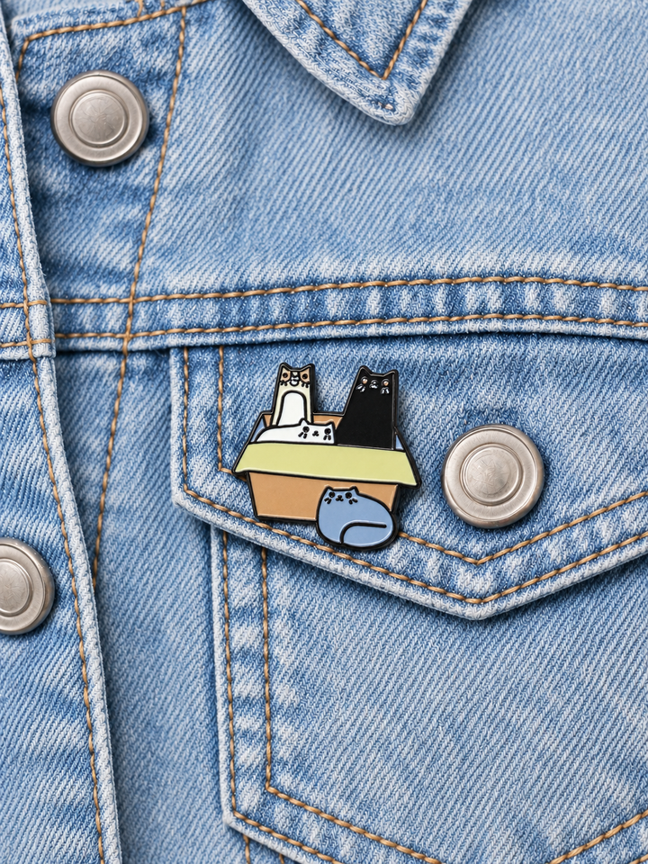 Cats in Cardboard Box Pin displayed on a blue denim jacket beside a metal button
