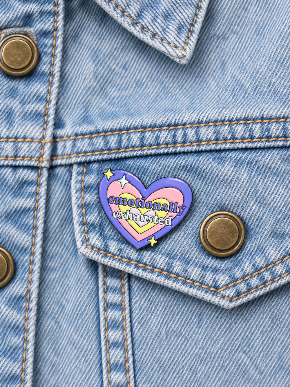 Emotionally Exhausted Heart Pin displayed on a light-wash denim jacket beside a metal button