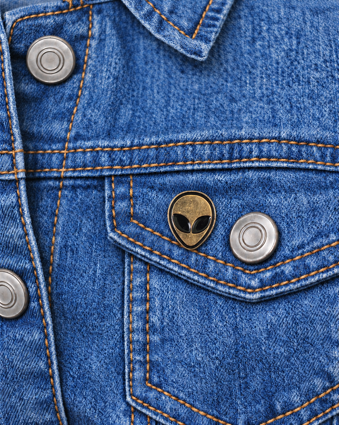 Gold Alien Head Pin displayed on a light-wash denim jacket beside a metal button