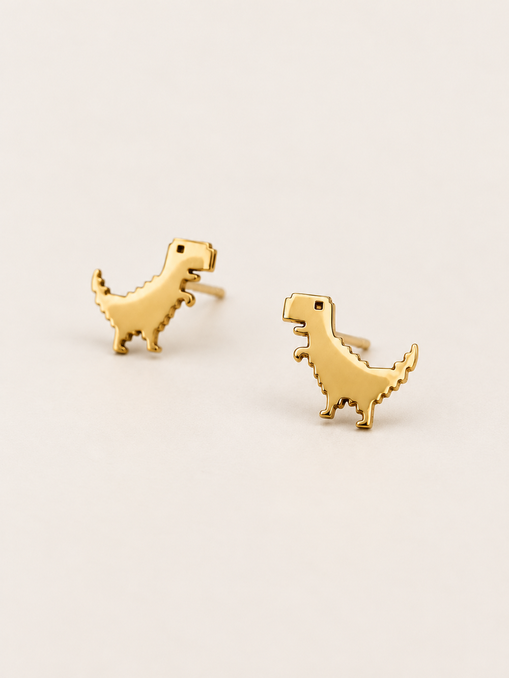 Gold T-Rex Stud Earrings shown angled on a warm neutral background