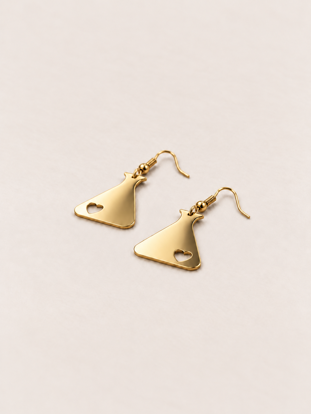 Gold Heart Flask Drop Earrings lying flat at an angle on a warm neutral background
