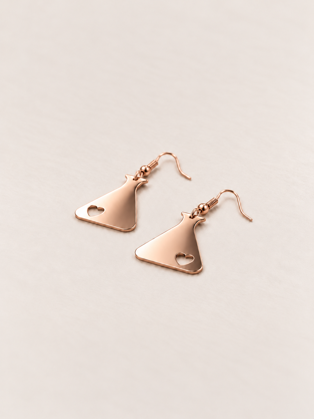 Rose Gold Heart Flask Drop Earrings lying flat at an angle on a warm neutral background
