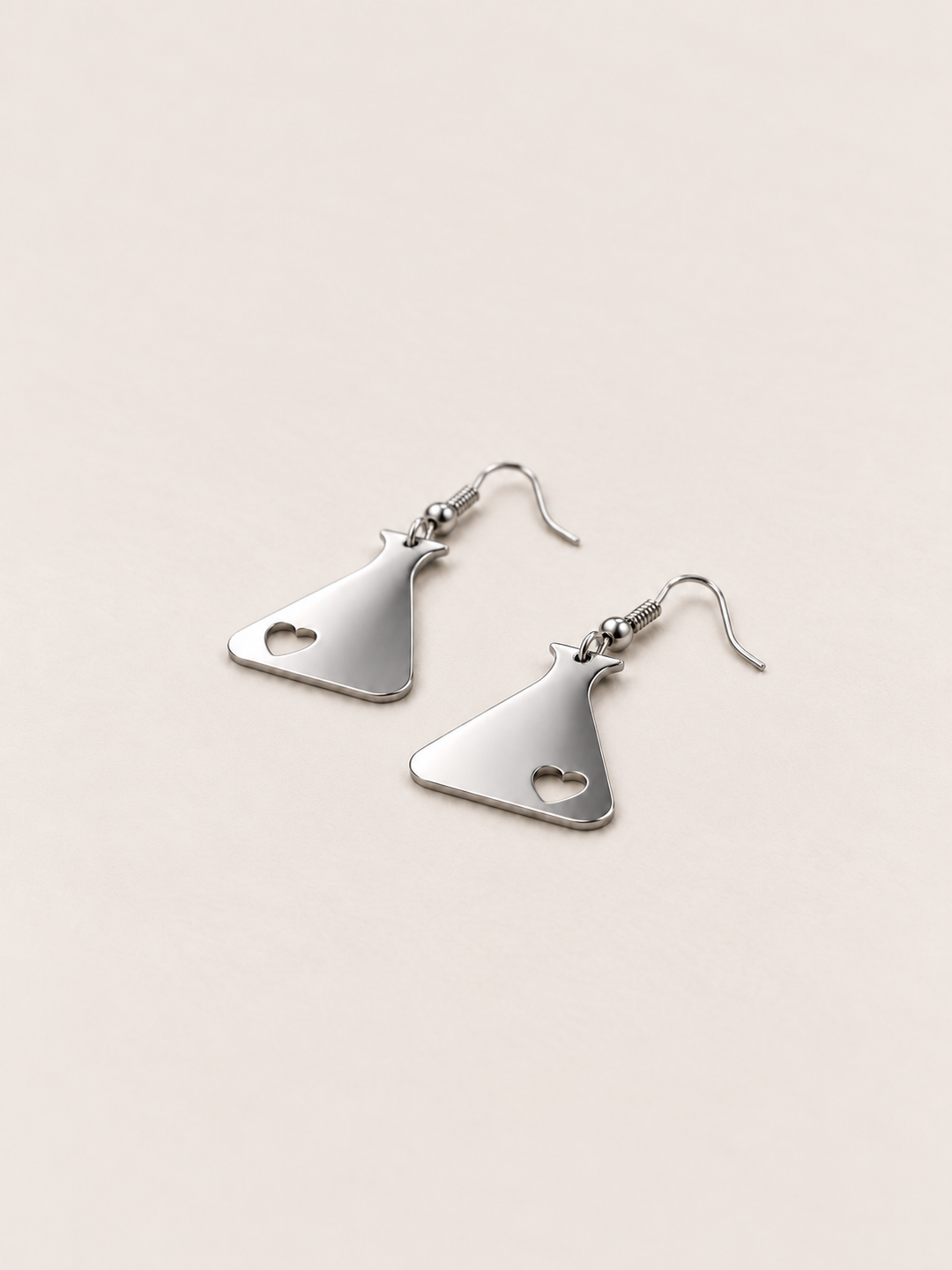 Silver Heart Flask Drop Earrings lying flat at an angle on a warm neutral background