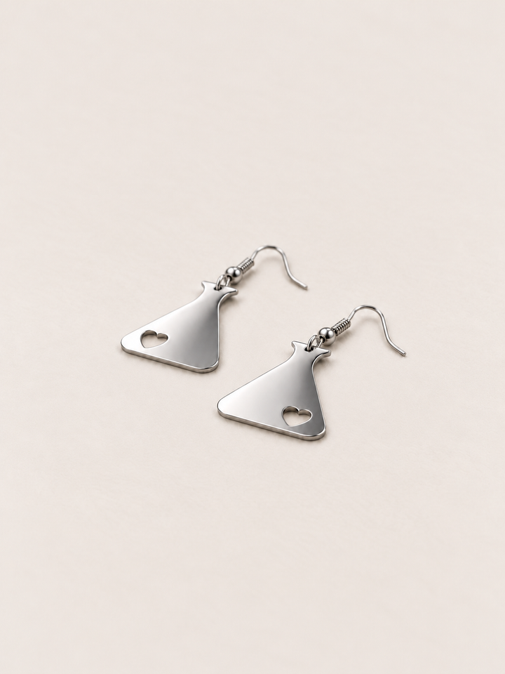 Silver Heart Flask Drop Earrings lying flat at an angle on a warm neutral background