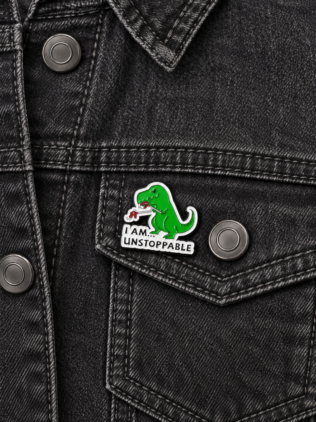I Am Unstoppable T-Rex Pin displayed on a charcoal denim jacket beside a metal button