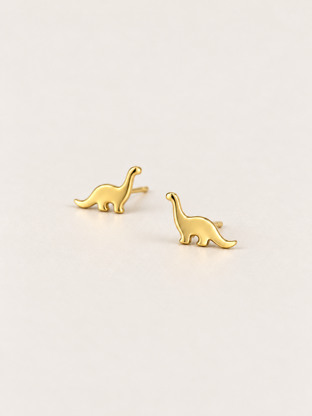 Long Neck Dinosaur Stud Earrings in gold shown angled on a warm neutral background