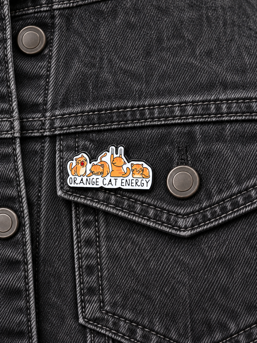 Orange Cat Energy Pin displayed on a blue denim jacket beside a metal button