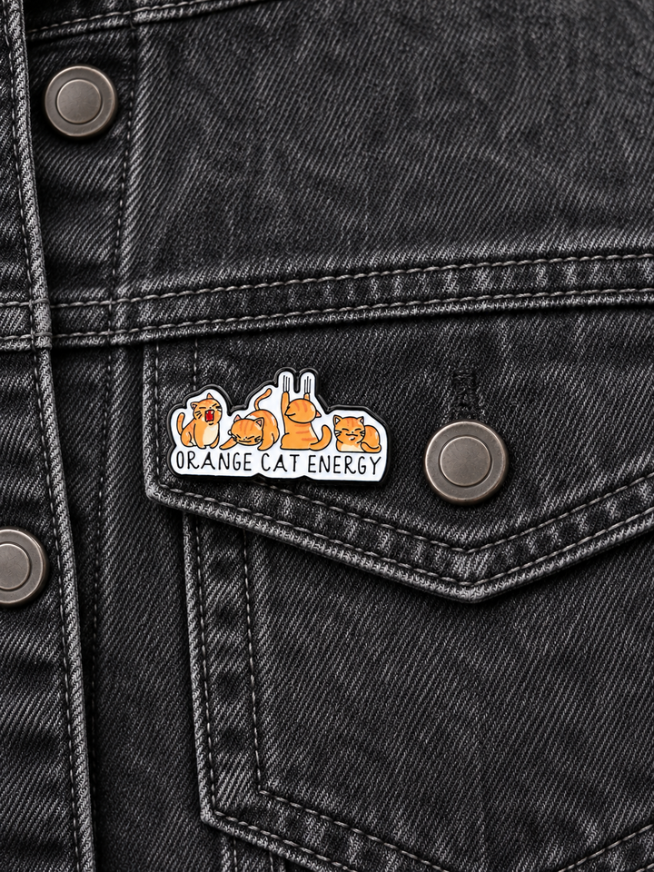 Orange Cat Energy Pin displayed on a blue denim jacket beside a metal button