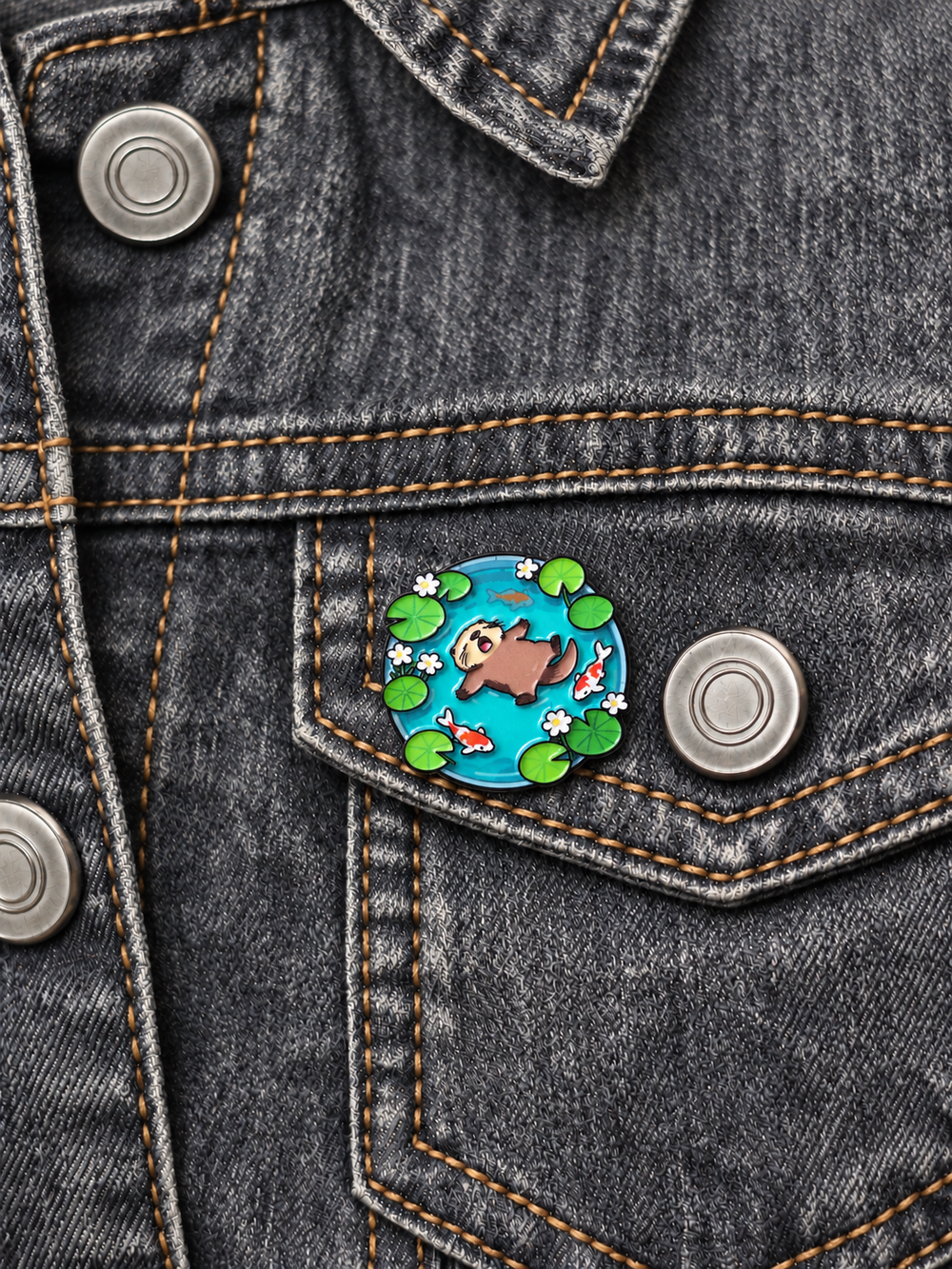 Otter Floating in Koi Pond Pin displayed on a faded gray denim jacket beside a metal button