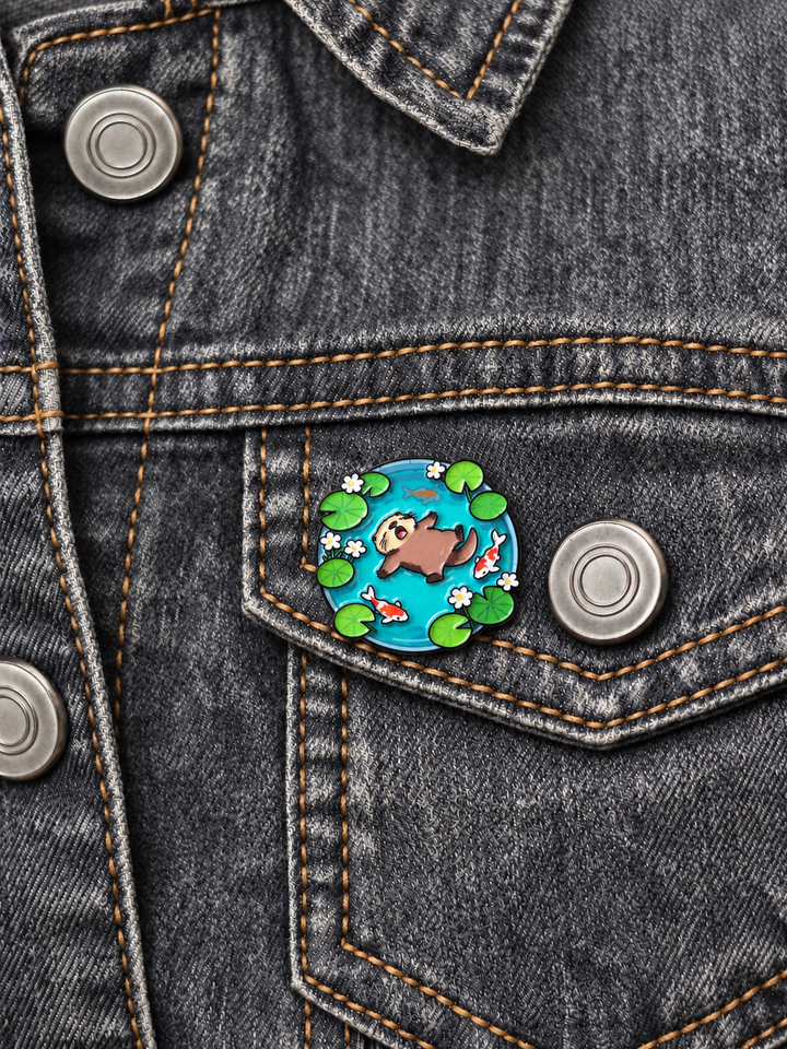 Otter Floating in Koi Pond Pin displayed on a faded gray denim jacket beside a metal button