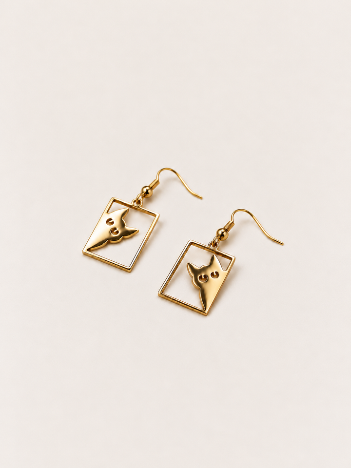 Gold Peeking Cat Rectangle Drop Earrings lying flat at an angle on a warm neutral background