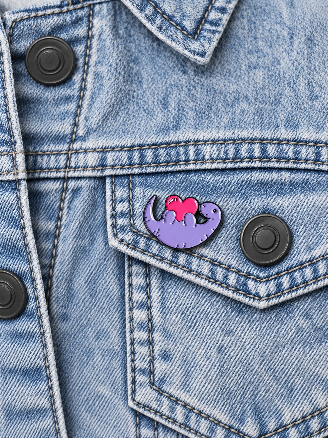 Purple Dinosaur Heart Pin displayed on an acid-wash denim jacket beside a metal button