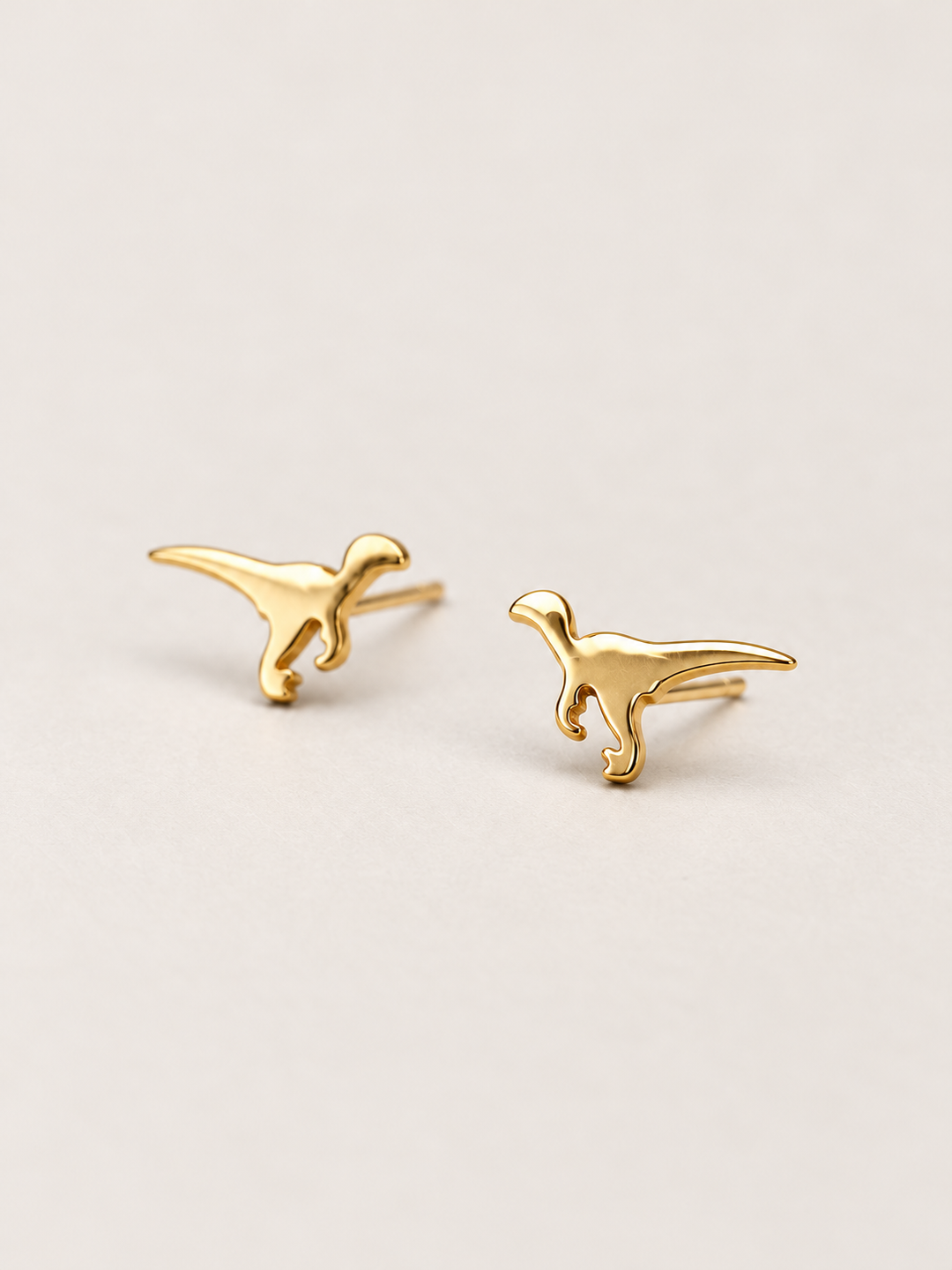 Gold Raptor Dinosaur Stud Earrings shown angled on a warm neutral background