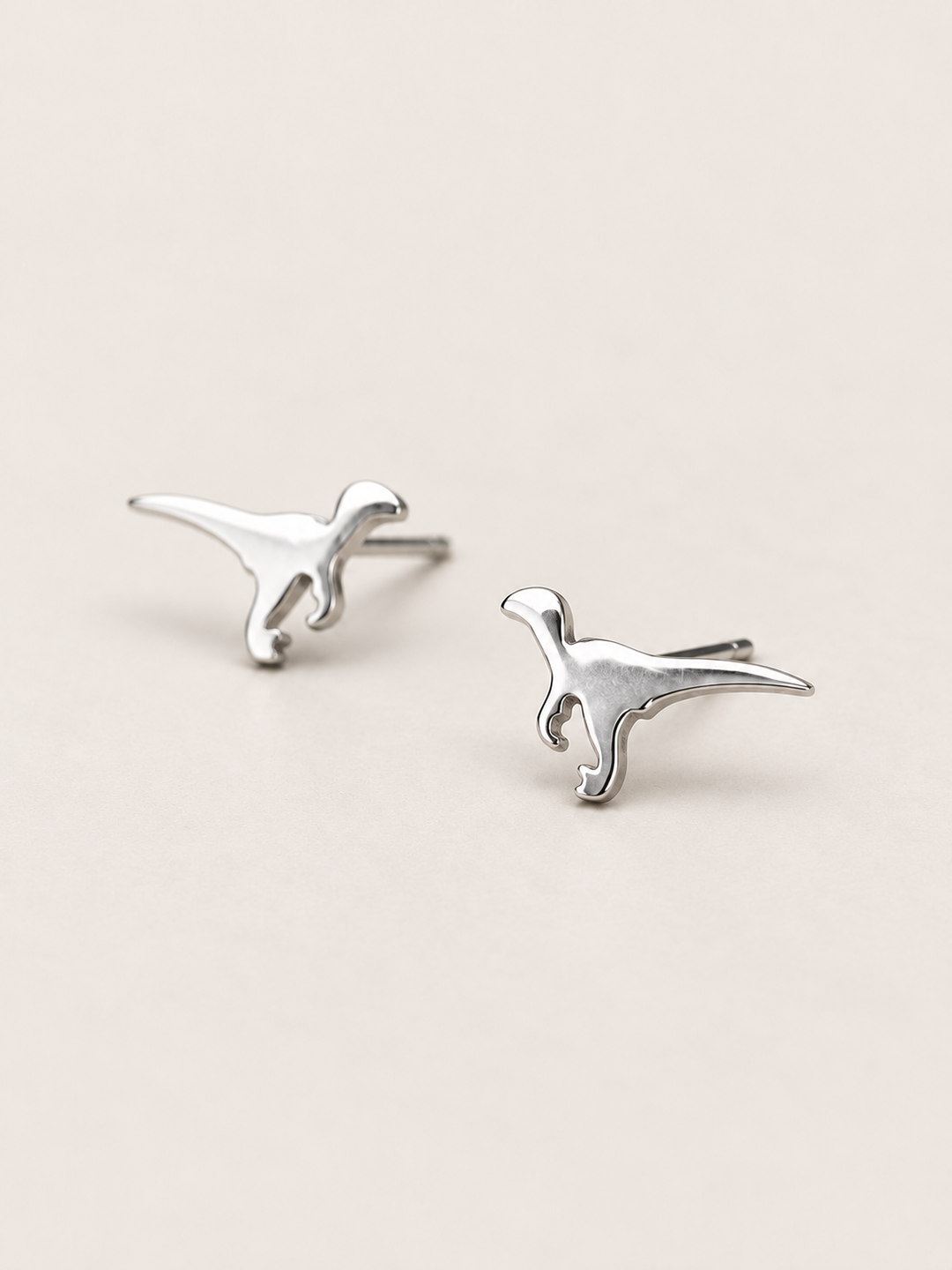 Silver Raptor Dinosaur Stud Earrings shown angled on a warm neutral background