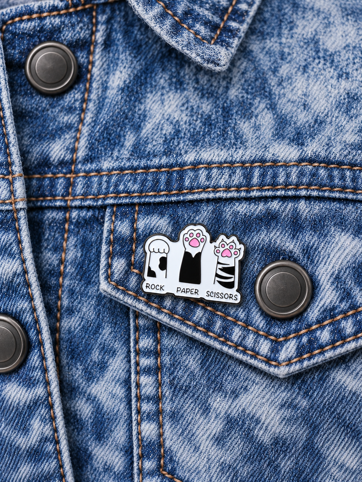 Rock Paper Scissors Cat Paws Pin displayed on an acid-wash denim jacket beside a gunmetal button