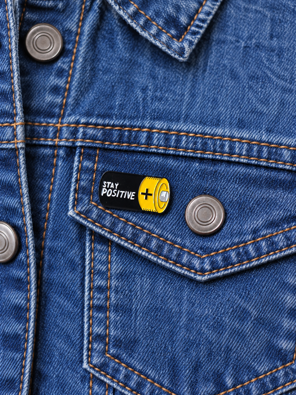 Stay Positive Battery Pin displayed on a stonewash denim jacket beside a metal button
