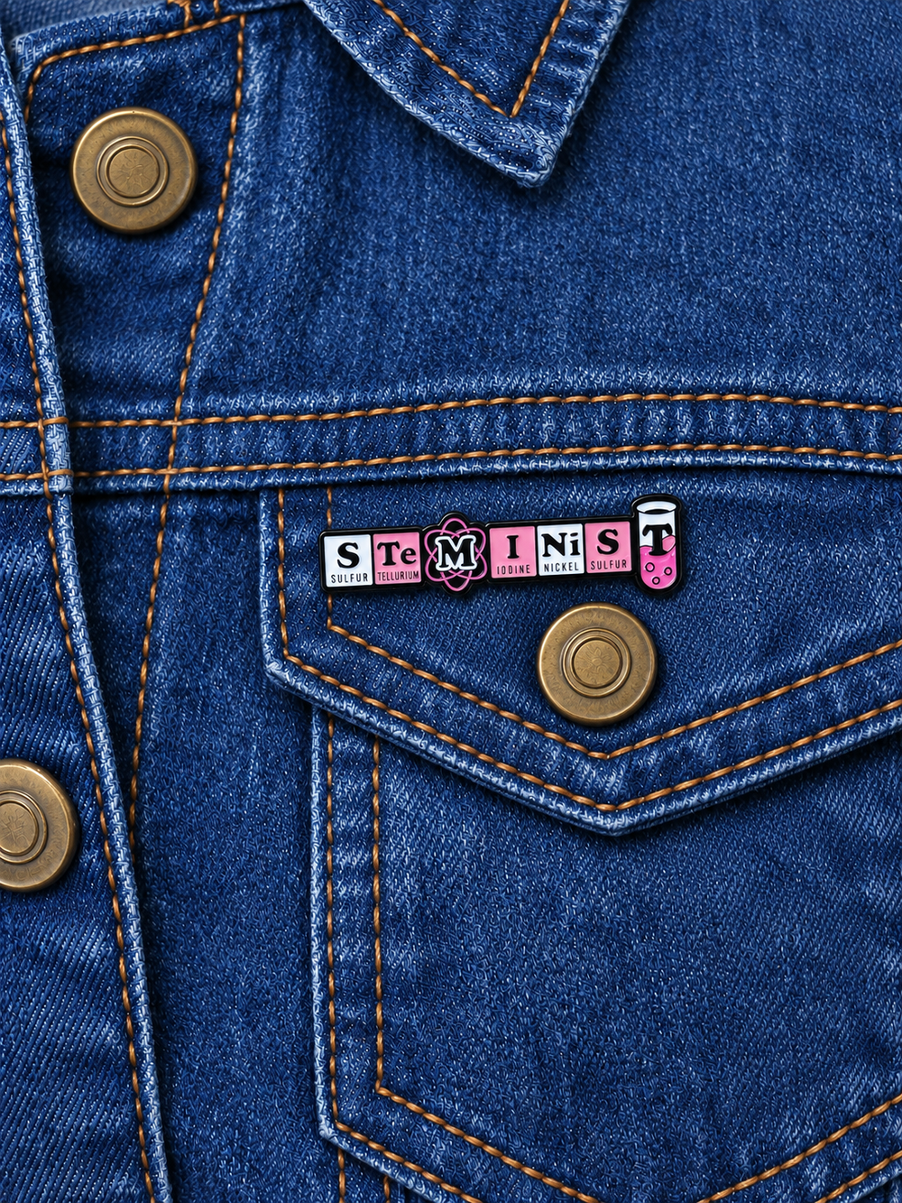 STEMinist Science Pin displayed on dark indigo denim beside a metal button