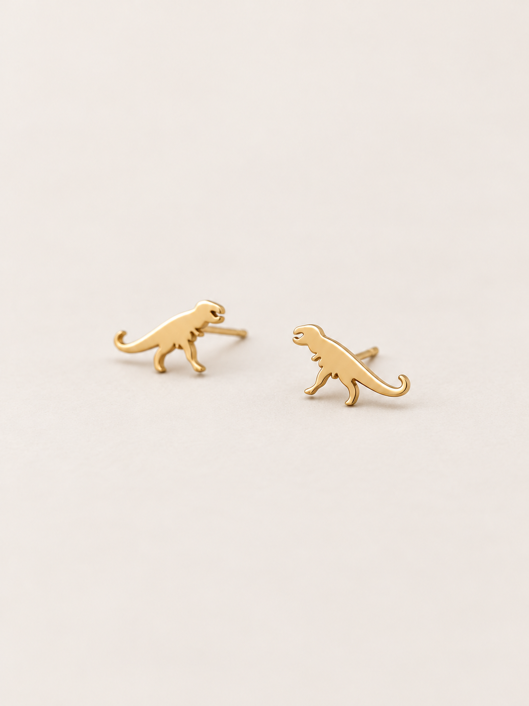 Gold T-Rex Dinosaur Stud Earrings shown angled on a warm neutral background