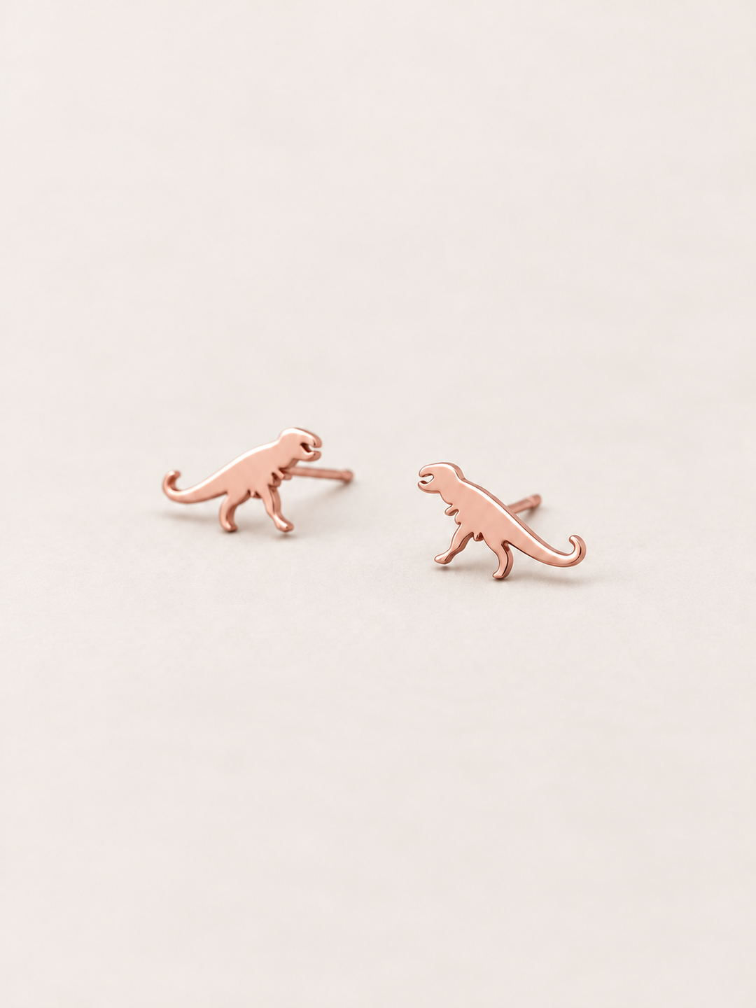 Rose Gold T-Rex Dinosaur Stud Earrings shown angled on a warm neutral background