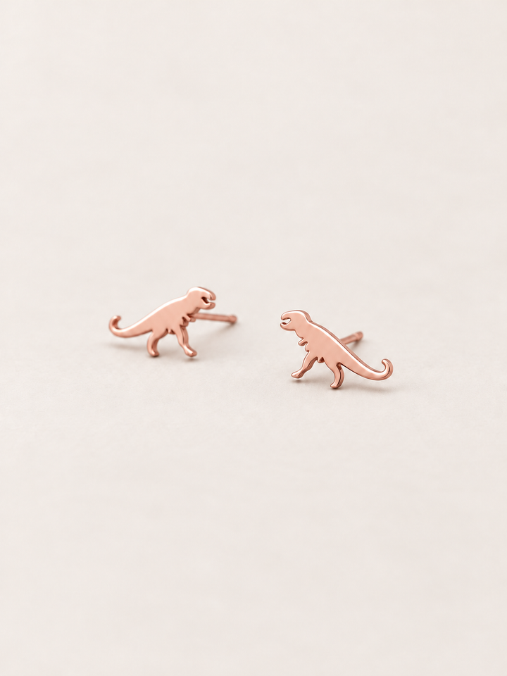 Rose Gold T-Rex Dinosaur Stud Earrings shown angled on a warm neutral background