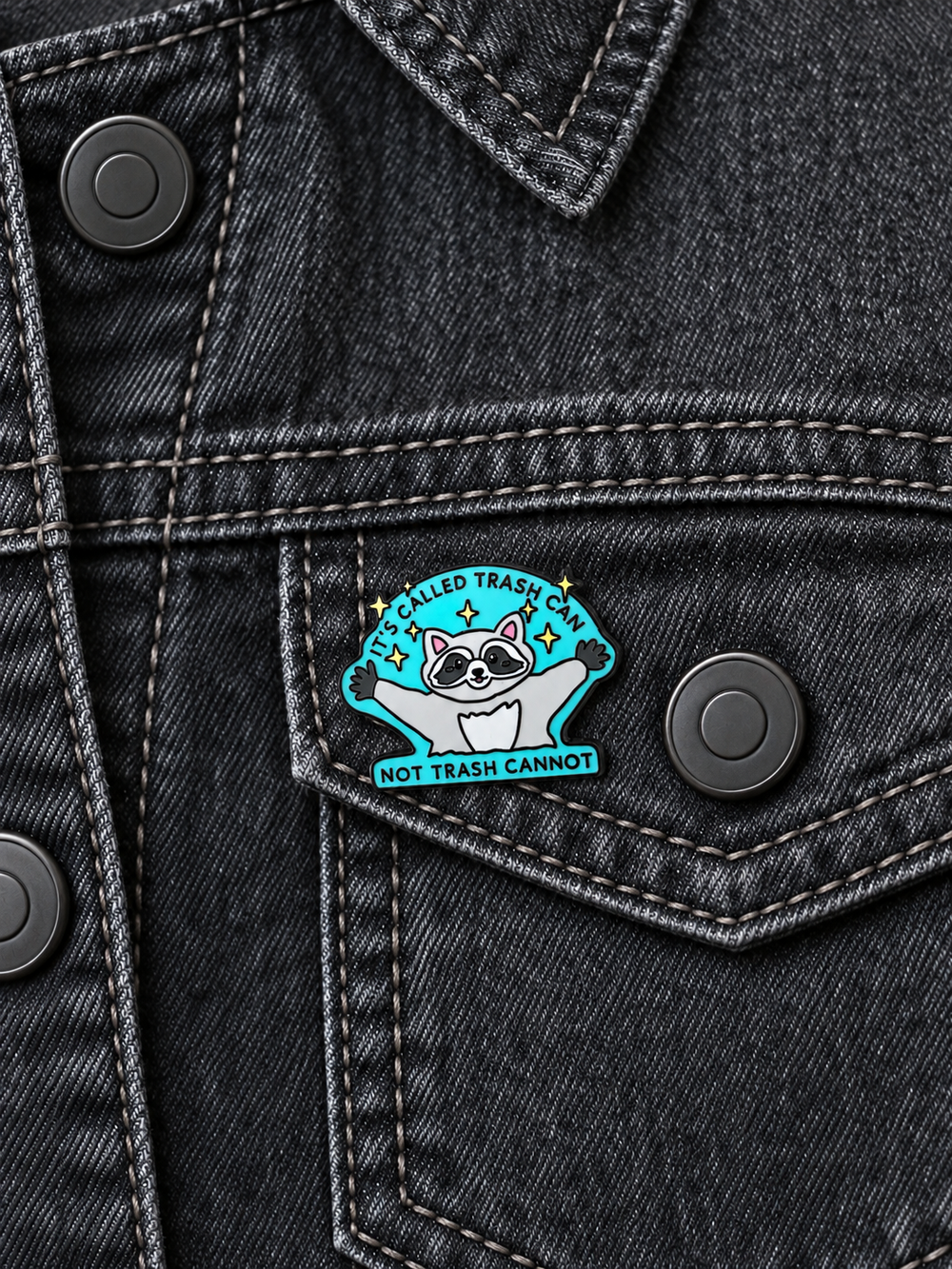 Trash Can Raccoon Pin displayed on a charcoal denim jacket beside a metal button