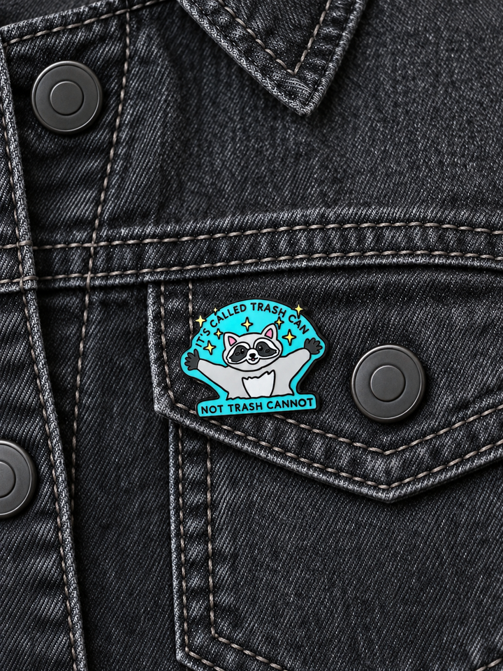 Trash Can Raccoon Pin displayed on a charcoal denim jacket beside a metal button
